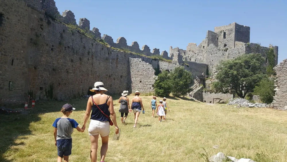 Groupe de familles et enfants explorant la cour intérieure herbeuse et les remparts historiques du château de Puilaurens sous un ciel bleu.