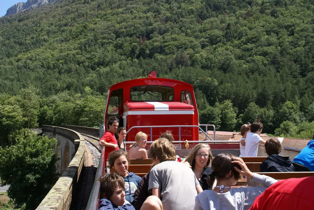 Enfants et adultes profitant d'un voyage à bord du wagon ouvert du Train Rouge traversant un viaduc au cœur des paysages naturels de la vallée